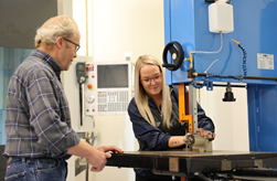 A man and a woman are engaged in using a bandsaw in a workshop. The woman operates the machine while the man observes.