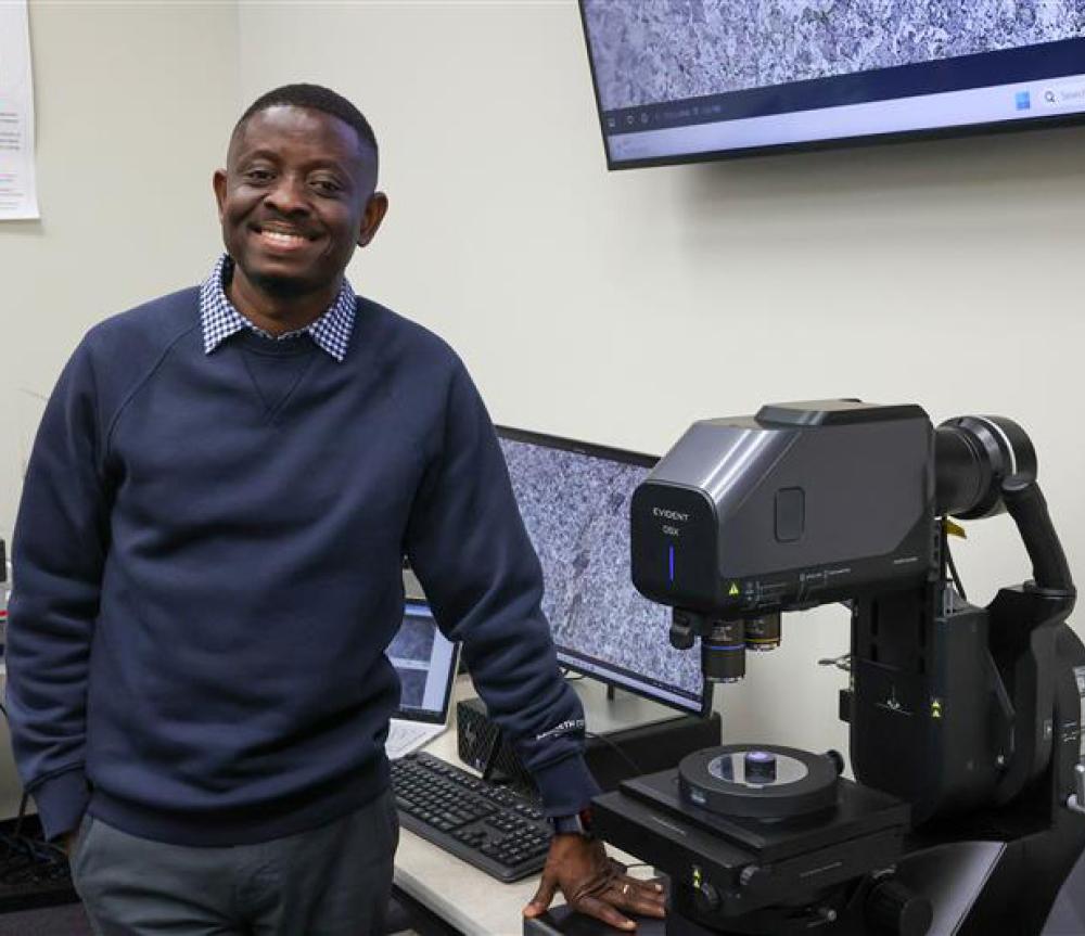 Tabiri in a sweater in front of research equipment. Smile on his face. 