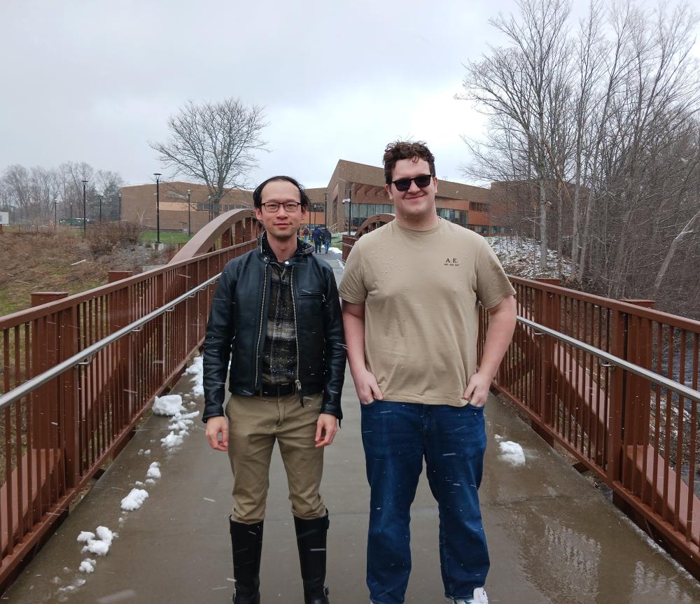 Dr. Leung in leather jacket and Student Research Seth Walther in t-shirt and jeans standing on bridge on campus on a late winter day.