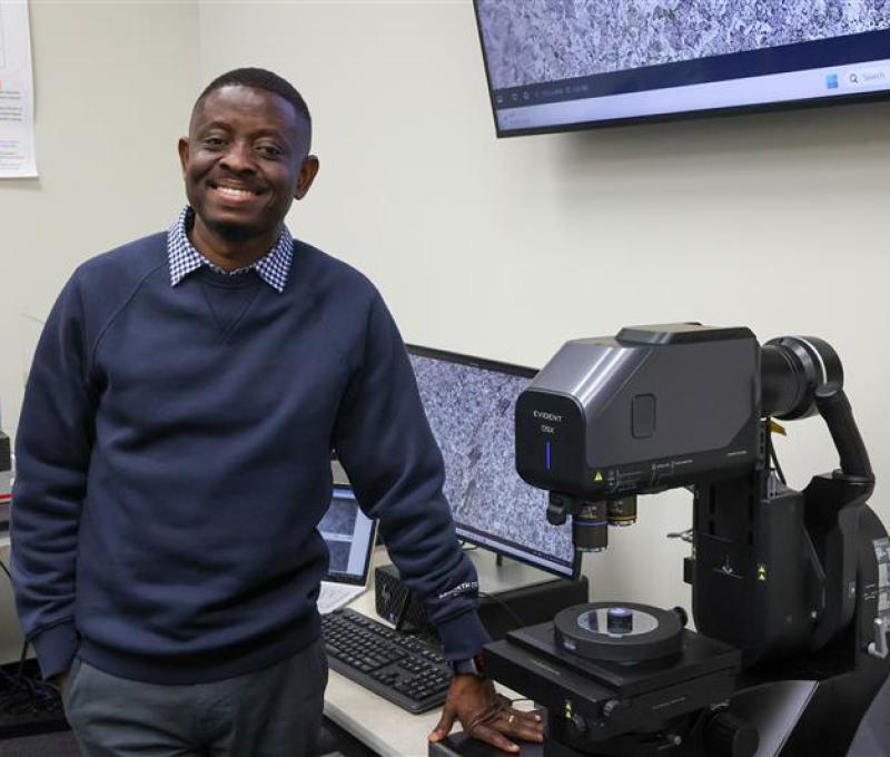 Tabiri in a sweater in front of research equipment. Smile on his face. 