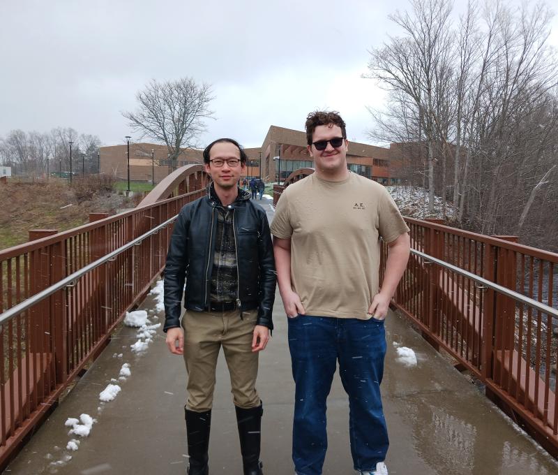 Dr. Leung in leather jacket and Student Research Seth Walther in t-shirt and jeans standing on bridge on campus on a late winter day.