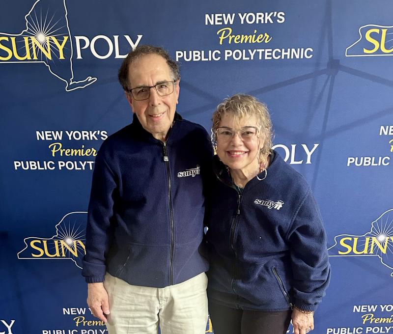 Ron and Pat Sarner wearing matching pullovers. SUNY Poly backdrop in background.